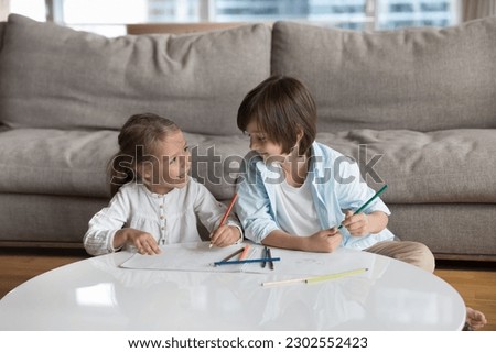 Cheerful little sibling children enjoying creative hobby, leisure, daycare activity at home together, chatting, talking, drawing with pencils, discussing colorful graphic doodles