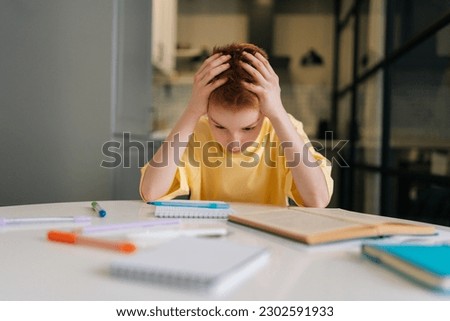 Portrait of exhausted pupil boy tired from studying holding head head with hands sitting at desk with paper copybook, looking down. Frustrated child schoolboy doing homework at home. Royalty-Free Stock Photo #2302591933