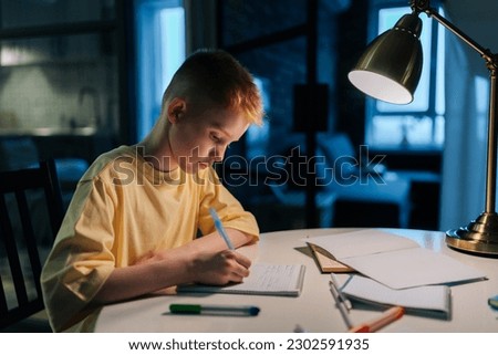 Side view of focused schoolboy studying at home doing homework sitting at table under light of lamp. Smart redhead pupil boy writing in exercise book at night in dark with lighting lamp. Royalty-Free Stock Photo #2302591935