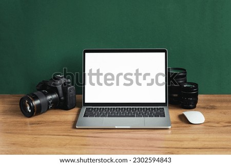 Modern photographer workplace with computer, DSLR camera and lenses. Laptop with the blank white screen on the wooden office table against dark green wall. Mockup for photo or videomaker advertisement
