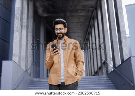 Successful young Hindu man walking outside office building, engineer software developer programmer smiling and happy using test app on phone, happy satisfied with result. Royalty-Free Stock Photo #2302598915