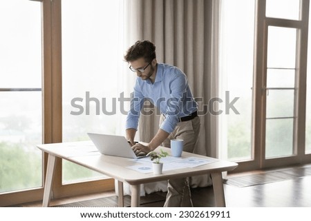 Serious young small business owner man working at laptop from home bending over workplace table with paper marketing, financial reports. Freelancer, entrepreneur, company leader candid shot