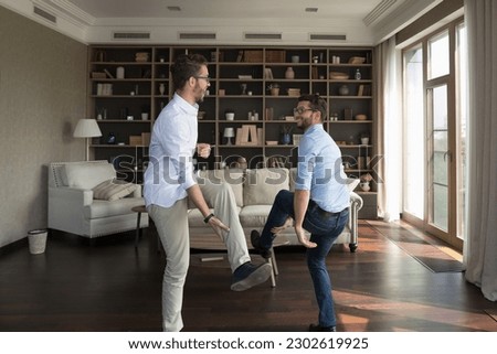 Two happy cheerful young adult twin brothers having fun at home, dancing together in living room, enjoying family party, active leisure, meeting, hopping, laughing