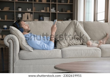 Peaceful millennial young man in big wireless headphones relaxing on comfortable sofa, listening to ambient music for meditation, using playlist on Internet, enjoying silent leisure, break at home Royalty-Free Stock Photo #2302619951