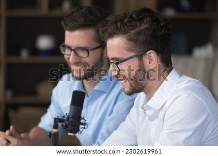 Happy positive young adult twin brothers broadcasting on air, speaking at studio microphone, recording podcast together, using sound equipment for Internet communication