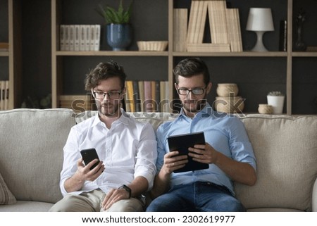 Serious twin brother working on family startup from home, doing freelance job, using mobile phone, tablet computer for online business communication, sitting on home couch together Royalty-Free Stock Photo #2302619977