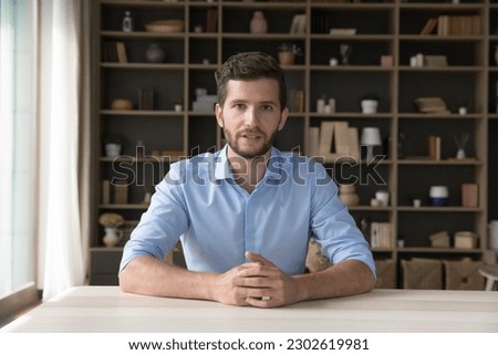 Serious handsome young online teacher man giving webinar on Internet, sitting at table, speaking, looking at camera with home interior in background. Business man, coach, blogger head shot portrait