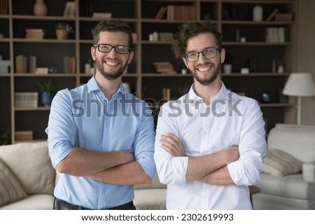Positive confident family business partner men portrait. Happy handsome young adult twin brothers in glasses and office shirts posing together at home with arms crossed, looking at camera, smiling