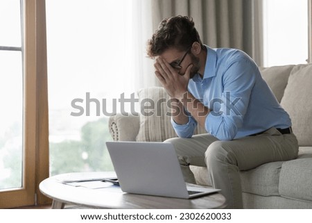 Stressed unhappy freelance business man sitting on home sofa at laptop, touching face, leaning head on hands in despair, thinking over overspending, bankruptcy, work problems Royalty-Free Stock Photo #2302620033