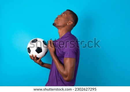 young man wearing purple T-shirt over blue studio background excited and glad to achieve victory, clenches fists, screams in excitement with closed eyes,successful person.
