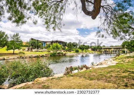 A visitor center on the other side of a tranquil river in San Angelo, Texas, surrounded by lush grass and trees with wispy clouds dotting a bright blue sky. Royalty-Free Stock Photo #2302732377