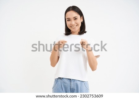 Portrait of cute smiling korean girl, 25 years, points at camera, invites you, congratulates, stands over white background.