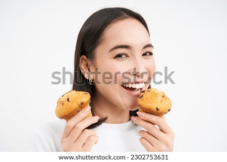 Image of happy korean girl who loves pastry, smiling and eating cupcakes, isolated on white background.