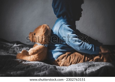 A sad little boy sits back to back with a teddy bear. Lonely depressed child in a room with a teddy bear. Post-traumatic disorder in a child Royalty-Free Stock Photo #2302763151