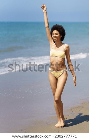 Full body of cheerful young African American female, traveler with dark curly hair in trendy bikini, smiling and looking away while walking on sandy beach with raised arm in sunlight