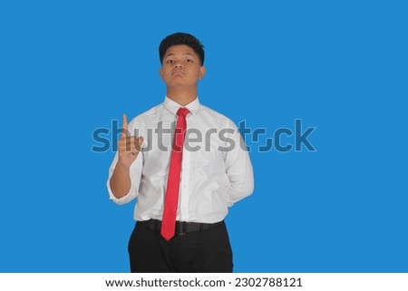 Asian male party cadre in tie pointing up with number one finger smiling confident and happy for election campaign isolated on blue background.