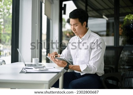 Smart and focused millennial Asian businessman or male boss using his digital tablet at his desk in the office.