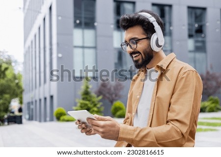 A young Inus man walks around the city in headphones, a businessman in a shirt watches an online video stream using a tablet computer, a programmer in a shirt outside an office building. Royalty-Free Stock Photo #2302816615