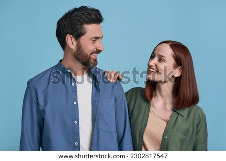 Portrait of smiling romantic couple looking each other, isolated on blue background. Handsome bearded man and beautiful woman standing together indoors. Love, relationship concept