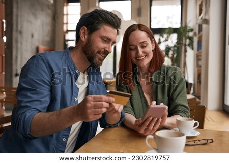 Smiling couple of friends holding credit card, using mobile phone shopping online, drinking coffee  sitting in modern cafe. Successful freelancers receive payment                               