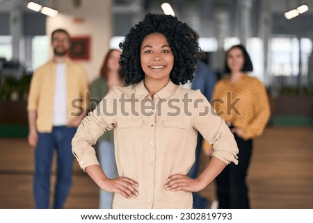 Portrait of smiling successful African American manager, businesswoman looking at camera standing in office with creative team on background. Successful business, career                              