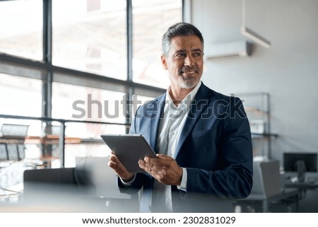 Happy middle aged business man ceo wearing suit standing in office using digital tablet. Smiling mature businessman professional executive manager looking away working on tech device. Authentic shot