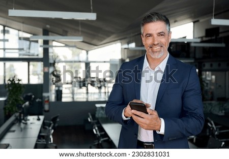 Happy mid aged business man ceo standing in office using mobile phone. Mature businessman professional executive manager digital crypto investor holding smartphone satisfied with corporate solution.