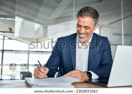 Happy satisfied middle aged professional business man executive ceo manager, lawyer wearing suit sitting at desk signing law document writing signature making legal agreement corporate deal in office. Royalty-Free Stock Photo #2302831061
