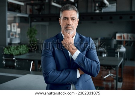 Serious confident mid aged business man, thoughtful doubtful company ceo executive wearing blue suit standing in office holding hand in chin looking at camera thinking, making decision, feeling doubt.