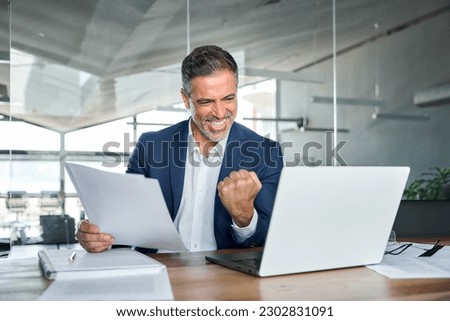 Happy mature older business man ceo wearing suit celebrating success at work in office holding papers looking at laptop rejoicing company growth, goals achievement good results screaming yes. Royalty-Free Stock Photo #2302831091