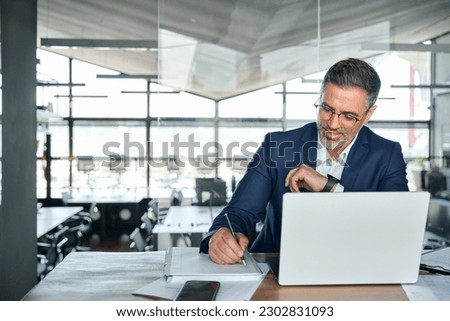 Middle aged smiling professional business man global company executive ceo manager or lawyer wearing suit sitting at desk in modern office working on laptop computer and writing notes, copy space. Royalty-Free Stock Photo #2302831093