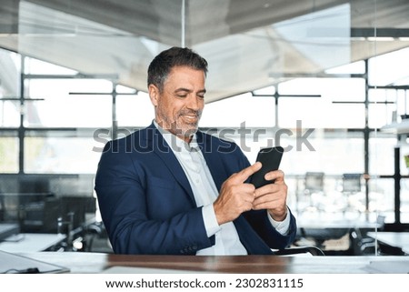 Happy mid aged business man ceo executive, mature financial manager investor wearing suit sitting in modern office holding smartphone using mobile phone managing digital tech transactions at work.