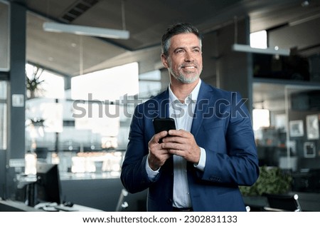 Smiling happy confident mid aged male company ceo executive wearing suit holding cellphone standing in office using business mobile apps technology financial online solutions on cell phone. Royalty-Free Stock Photo #2302831133