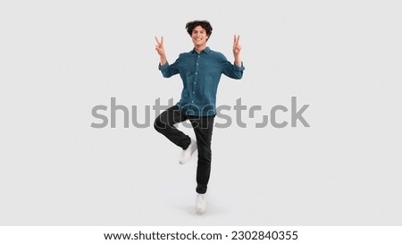 Victory. Funny Young Brunette Man Gesturing V Sign Smiling At Camera Posing Standing On One Leg Over Light Gray Background In Studio. Cheerful Guy Showing Peace Symbol With Both Hands. Panorama
