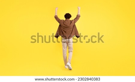 Victory Celebration. Rear View Of Young Man Raising Arms And Shaking Fists In Triumph, Embracing Success And Motivation Standing Back To Camera On Yellow Background. Full Length Shot, Panorama Royalty-Free Stock Photo #2302840383
