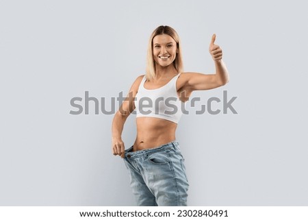 Effective diet, slimming concept. Happy cheerful beautiful blonde middle aged woman wearing big jeans after weight loss on grey studio background, showing thumb up at camera, copy space Royalty-Free Stock Photo #2302840491