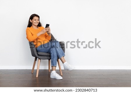 Cheerful positive attractive young indian woman wearing casual outfit sitting in armchair at home, using cell phone, chatting with guys on dating app, shopping online, scrolling, copy space Royalty-Free Stock Photo #2302840713
