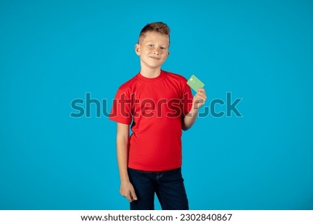 Cheerful Preteen Caucasian Boy Holding Credit Card In Hand And Looking At Camera, Smiling Happy Male Child Recommending Bank Services While Standing Isolated Over Blue Background, Copy Space
