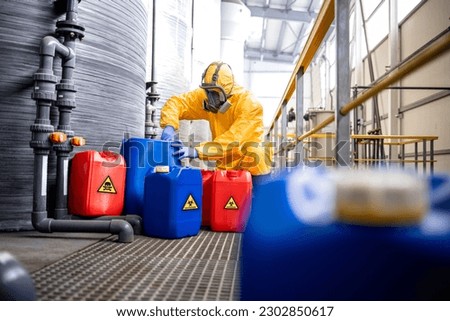 Factory worker in hazmat suit and gas mask filling plastic container with chemical and hazardous waste. Chemicals factory interior with acid storage tanks. Royalty-Free Stock Photo #2302850617