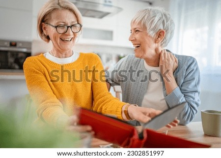 Senior mature women friends or sisters looking family photos album two caucasian females sit at home happy smile while revoke memories real people copy space