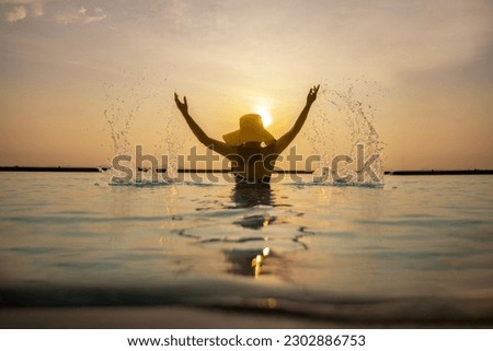 Back side view of Woman in bikini and yellow hat in infinity swimming pool with drops of water splashed by a young woman and Golden sky with sun and calm ocean at sunset.