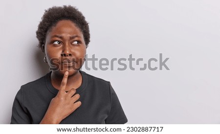 Horizontal shot of serious pensive dark skinned woman keeps finger on chin considers something being deep in thoughts dressed in casual black t shirt isolated over white background copy space for text