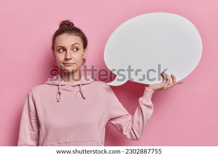 Thoughtful pretty young woman with dark hair gathered in bun holds abstract cloud bubble for thoughts and words dressed in casual sweatshirt isolated over pink background. Communication concept