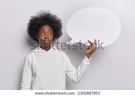 Thoughtful doubtful woman with Afro hair holds blank speech bubble thinks what information to write there wears casual sweatshirt stands against white background. People idea and contemplation concept