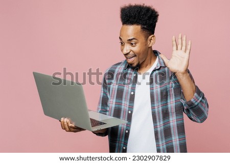 Young smiling happy man of African American ethnicity 20s he wear blue shirt hold use work on laptop pc computer waving hand talk get video call isolated on plain pastel light pink background studio