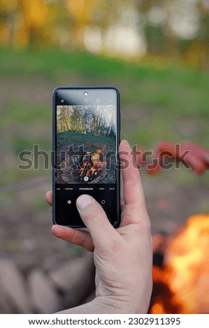 Smartphone in male hand taking picture of roasting sausages skewered on a grill fork over an open campfire. Grilling sausages for dinner at camping or in the garden. Taking picture by smartphone