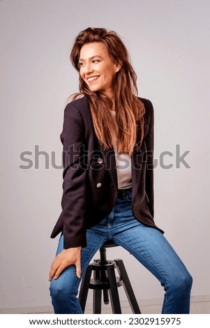 Studio shot of a beautiful smiling woman sitting at isolated grey background. Happy woman wearing black blazer with white shirt and blue jeans. Copy space. 
