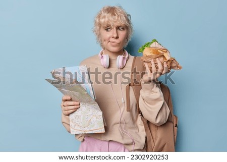Female traveler holds paper map searches for route eats sandwich purses lips has dimpes in cheeks dressed in sweatshirt poses with backpack has adventurous trip isolated over blue background.