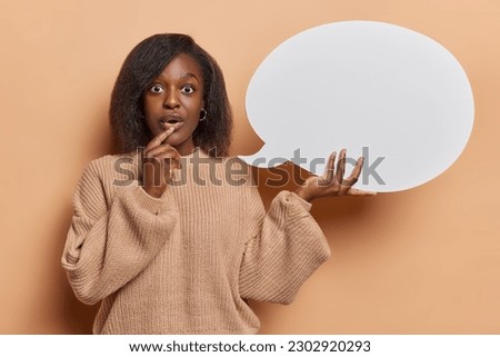 Dont miss this chance. Stunned black woman with curly hair holds speech bubble stands amazed keeps mouth opened dressed in knitted sweater isolated over brown background. Place your promo here