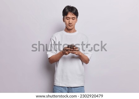 Horizontal shot of handsome Asian man types something on smartphone chats in messenger checks social networks has serious expression dressed in casual t shirt isolated over white background.